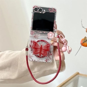 Puede incluir: Funda transparente para teléfono con diseño de mariposa roja y correa roja a juego. La funda tiene un estampado floral y un pequeño adorno de flor rosa. El teléfono está abierto, revelando una pantalla negra. La correa incluye un charm rosa.