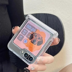 Puede incluir: Funda protectora transparente para teléfono inteligente plegable. La pantalla del teléfono muestra un perro con gafas y un suéter, con la hora 00:28. La funda tiene un marco plateado y una parte trasera transparente.