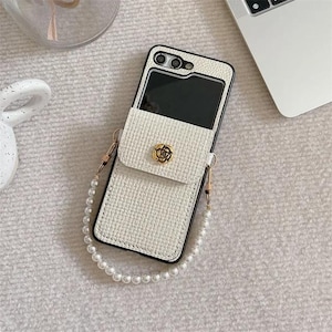 Puede incluir: Funda de teléfono blanca texturizada con una pequeña solapa tipo bolso y una correa de perlas. La solapa tiene un emblema de flor dorada. La funda del teléfono es negra en los bordes y tiene una abertura para la cámara.