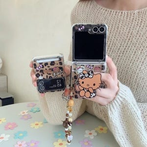 Puede incluir: Funda transparente para teléfono con diseño de estampado de leopardo y un gráfico de Hello Kitty. La funda se muestra abierta y cerrada, con un charm decorativo. La funda está diseñada para un teléfono plegable.