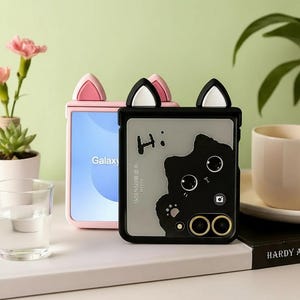 Może przedstawiać: Dwa etui na telefon z motywem kocich uszu. Jedno jest różowe z niebieskim ekranem z napisem "Galaxy". Drugie jest czarne z sylwetką kota i napisem "Hi:" na etui. Etui znajdują się na białej powierzchni.