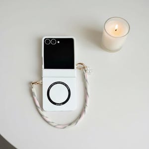 May include: A white foldable phone case with a black screen and a circular design. The case has a matching strap with pink, grey, and white accents, and a pearl charm. A lit candle in a glass is also present.