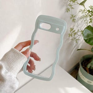 May include: A clear phone case with a wavy, light blue border. The case has cutouts for the camera and other phone features. The case is held by a person. The background is a light color with a plant in a pot.