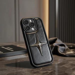 May include: Black leather phone case featuring a silver starburst emblem. The case has a raised border around the camera and a textured leather finish. The starburst is centrally placed on the back of the case.
