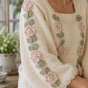 May include: A cream-colored crochet sweater featuring a square neckline and long sleeves. The garment is decorated with vertical rows of light pink crocheted flowers and sage green crocheted leaves applied to the front and along the sleeves.