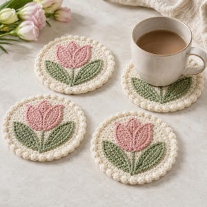 May include: Four handmade crochet coasters with a tulip design. Each coaster features a pink tulip with green leaves on a cream-colored background, surrounded by a scalloped edge. One coaster holds a beige mug with a brown liquid.