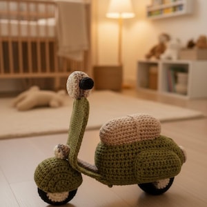 Può includere: Un giocattolo scooter all'uncinetto fatto a mano in verde oliva, beige e nero. Lo scooter ha un corpo arrotondato, ruote e manubrio. Il giocattolo è per bambini ed è posto su un pavimento in legno chiaro.