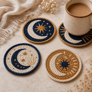 May include: Four handmade crochet coasters with celestial designs. Each coaster features a crescent moon and starburst pattern in navy blue, cream, and gold. One coaster has a mug of coffee on it. The coasters are round and about 4 inches in diameter.
