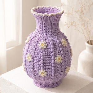 May include: A lavender crochet vase featuring a ribbed texture with vertical rows of small bobble stitches. The vase is decorated with several white and yellow daisy appliques and finished with a scalloped cream-colored trim at the top rim.