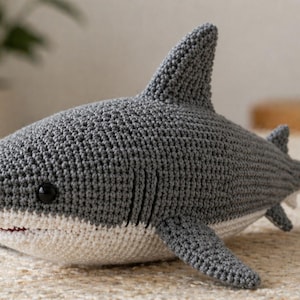 May include: A crocheted shark plushie in shades of gray and white. The shark has a detailed crochet texture, black eyes, and a white belly. The fins and tail are also gray. This handmade item is a soft toy.