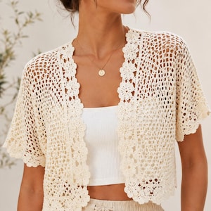 May include: Cream-colored crochet cardigan with short sleeves and a floral trim. The open-front design features a delicate, open-weave pattern. The cardigan is worn over a white top and paired with beige pants.