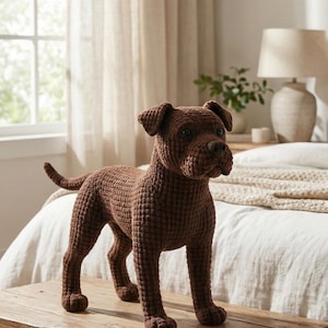 May include: A brown crochet dog figurine stands on a wooden surface. The dog has a textured appearance, with detailed features including a nose, ears, and tail. The figurine is a decorative item, likely for home decor.