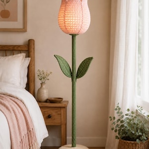 May include: A crochet floor lamp shaped like a tulip. The lamp features a pink flower head, green stem and leaves, and a cream-colored base. The lamp is placed in a bedroom setting, next to a bed with a pink blanket.