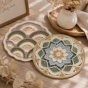 May include: Two handmade crochet coasters. One has a fan design in cream, olive green, and tan. The other features a floral pattern in cream, light blue, and olive green. Both are on a wooden surface.