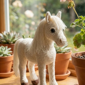 Pode incluir: Figura de cavalo de crochê branco sujo, com crina e cauda esvoaçantes. O cavalo tem cascos castanhos escuros e olhos pretos. Está sobre uma superfície de madeira, com plantas em vasos ao fundo.