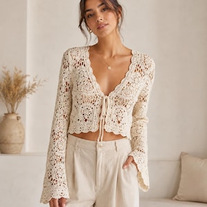May include: A cream-coloured long-sleeve crochet cardigan featuring an intricate floral pattern, a deep V-neckline, and a front tie closure. The garment has flared sleeves and a cropped hemline, showcasing detailed openwork stitching throughout the fabric.