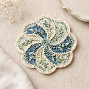 Könnte beinhalten: Ein handgefertigter, gehäkelter Untersetzer in floralem Design. Der Untersetzer hat ein Spiralmuster mit abwechselnden Abschnitten in Creme, Hellblau und Dunkelblau. Zarte grüne Blattakzente sind gleichmäßig verteilt. Der Untersetzer hat einen Durchmesser von etwa 15 cm.