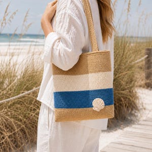 May include: A beige, white, and blue striped crochet tote bag with a seashell applique. The bag has a rectangular shape with a matching strap. The background shows a beach scene with sand and tall grass.
