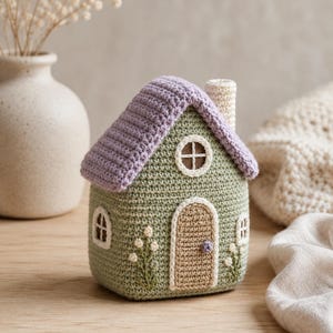 May include: A crocheted house figurine featuring a sage green body, a lavender roof, and a cream-colored chimney. The house has a tan arched door with a small purple knob, three white-trimmed windows, and embroidered white floral accents.