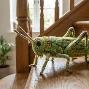 Könnte beinhalten: Eine handgefertigte Heuschrecke aus Häkelarbeit in Grün- und Gelbtönen mit braunen Akzenten. Die Heuschrecke steht auf einer Holztreppe und zeigt ihre detaillierte Textur und langen Fühler. Ein einzigartiges dekoratives Objekt.