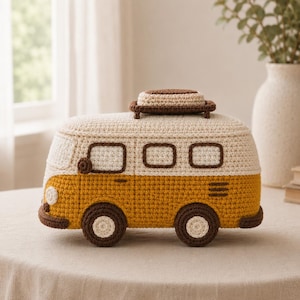 May include: A crocheted camper van in cream, mustard yellow, and brown. The van has brown wheels, windows, and a luggage rack on top. The vehicle is sitting on a neutral-colored surface.