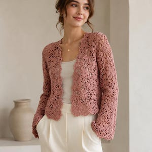 May include: A dusty rose crochet cardigan featuring an intricate floral pattern throughout the fabric, long sleeves, and a scalloped edge along the front opening. The garment is worn over a plain light-colored top and high-waisted cream trousers.