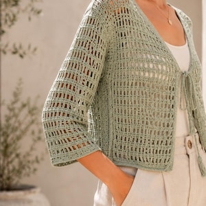May include: A light green, open-weave crochet cardigan with three-quarter sleeves. The cardigan has a V-neck and a tie closure at the front. The garment is worn over a white top and paired with beige pants.