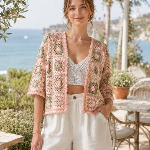 May include: A woman wearing a crochet cardigan with a floral pattern in pink, green, and cream colors. The cardigan has a scalloped edge and is paired with a white top and shorts. The background shows a coastal scene with the ocean and a sunny day.