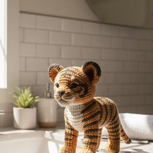 May include: A handmade crochet tiger cub in shades of orange, black, and white. The amigurumi tiger has a detailed striped pattern, small ears, and a sweet expression. The toy is standing on a countertop.