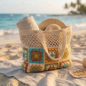 May include: A crocheted beach bag filled with a rolled-up towel and a straw hat sits on a sandy beach. The bag features a colorful patchwork design. A ball of yarn, a crochet hook, and a crocheted square are next to the bag.