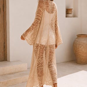 May include: A long, beige crochet cover-up with bell sleeves. The cover-up is worn over a white garment. The open weave design is visible. The model is wearing beige sandals. The background is a neutral color.