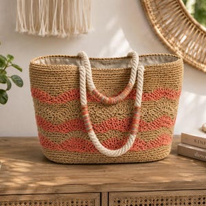 May include: A woven straw tote bag with coral-colored horizontal stripes. The bag has rope handles and a neutral-colored interior. The bag is sitting on a wooden surface.