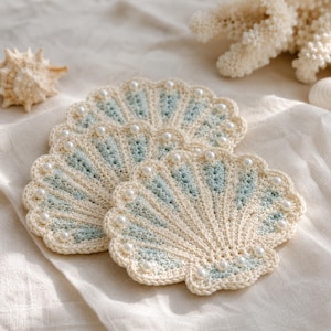 May include: A set of handmade crochet seashell coasters in cream, pale blue, and white, decorated with faux pearls. The coasters are displayed on cream fabric, with a seashell and coral in the background. Ideal for a coastal-themed home.