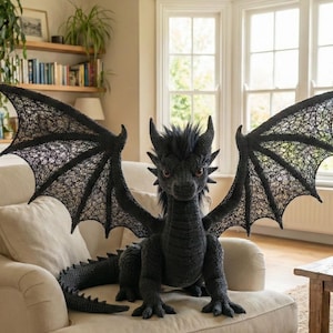 Puede incluir: Un gran dragón negro tejido a ganchillo con alas extendidas está sentado en un sofá beige. El dragón tiene detalles como cuernos, una melena puntiaguda y alas texturizadas. Las alas tienen un diseño delicado y calado. Los ojos del dragón son de color ámbar.