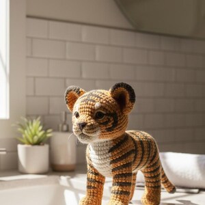 May include: A handmade crochet tiger cub in shades of orange, black, and white. The amigurumi tiger has a detailed striped pattern, small ears, and a sweet expression. The toy is standing on a countertop.