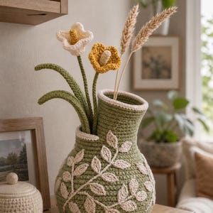 Op de afbeelding: Een handgemaakte gehaakte vaas in olijfgroen, versierd met crèmekleurige bladerpatronen. De vaas bevat gehaakte bloemen in wit en geel, samen met gedroogde tarwearen. De vaas staat op een houten oppervlak en toont een rustieke, handgemaakte esthetiek.