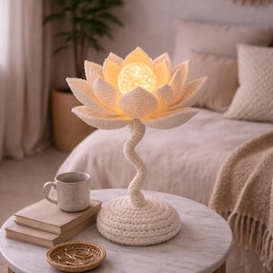 May include: A cream-colored crochet lotus flower lamp with a glowing center, set on a round, marble-topped table. The lamp's stem is a twisted design, and the base is a crocheted circle. A mug and books are on the table.