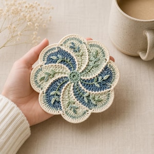 Könnte beinhalten: Ein handgefertigter, gehäkelter Untersetzer in floralem Design. Der Untersetzer hat ein Spiralmuster mit abwechselnden Abschnitten in Blau, Grün und Creme. Zarte blattartige Details sind in das Design eingearbeitet. Eine Tasse Kaffee steht in der Nähe.