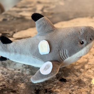 May include: A plush gray shark toy with black fins and a white belly. The shark has a light beige patch on its side and a small, round eye. The toy is made of soft, fuzzy material.