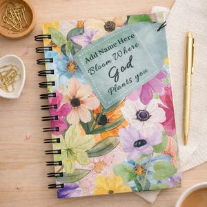Puede incluir: Un cuaderno con espiral con un diseño floral colorido. El cuaderno tiene una etiqueta que dice "Add Name Here Bloom Where God Plants you". También se ve un bolígrafo dorado y una taza de café.