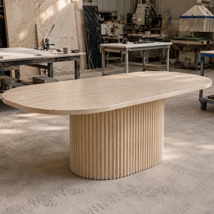 May include: An oval dining table with a smooth, light beige stone top and a cylindrical base featuring vertical fluted detailing. The table is constructed from a solid, textured material with a consistent neutral color throughout the entire piece.