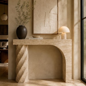 May include: A beige travertine console table with a spiral column leg and an arched side. On top sits a dark brown vase with green branches, a small bowl, a book titled Live Beautifully, and a mushroom-shaped lamp with a white glass shade.