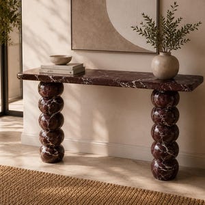 May include: A console table crafted from deep burgundy marble with white veining, featuring a rectangular top supported by two vertical columns composed of stacked, rounded marble spheres. The surface holds a small bowl and two stacked books.