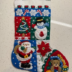 May include: Handmade Christmas stocking with a patchwork design. Features a snowman, Santa Claus, Christmas tree, and a teddy bear. The stocking is made of felt and decorated with sequins and beads. Includes a red loop for hanging.