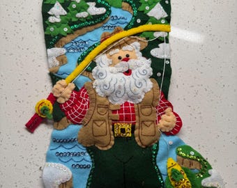 Op de afbeelding: Een kerstsok met de kerstman die aan het vissen is. De blauwe sok heeft een viltontwerp van de kerstman in een visvest, een rivier en een vis. De sok is versierd met pailletten en een rode lus om op te hangen.