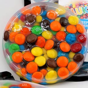 Puede incluir: Un recipiente transparente y redondo lleno de caramelos de colores. Los caramelos son de varios colores, incluyendo naranja, amarillo, verde, azul, rojo y marrón. Los caramelos están en una sustancia clara similar a un gel. Una etiqueta con la palabra "Luks" es visible.