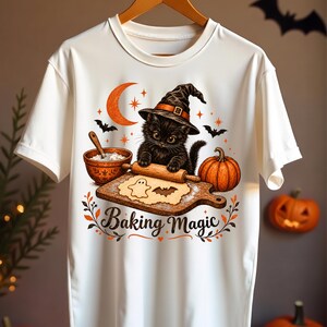 May include: A white t-shirt featuring a graphic of a black kitten wearing a witch hat, rolling dough on a wooden board with a ghost and bat-shaped cutout. The design includes a bowl, a pumpkin, a crescent moon, and the text Baking Magic.