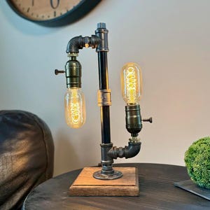May include: Industrial-style table lamp featuring a black pipe design with two Edison-style light bulbs. The lamp is mounted on a wooden base and has a vintage aesthetic. The lamp is approximately 12 inches tall.
