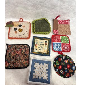 May include: A selection of handmade quilted pot holders in diverse shapes and patterns. Features a scarecrow, a green crocheted square, a Christmas-themed square, a paisley print, a square with the text "Keep Calm and Quilt On", a blue snowflake design, and a round holiday design.