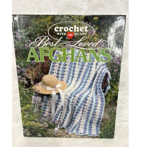 May include: A hardcover book titled Crochet With Heart Best Loved AFGHANS featuring a blue, white, and purple striped crocheted blanket draped over a chair with a straw hat with a white ribbon placed on top, set against a garden background.